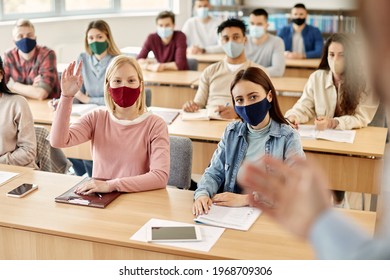 Large Group Of College Students Wearing Face Masks While Attending Lecture During Coronavirus Pandemic. Focus Is On Happy Student Raising Her Hand To Answer Teacher's Question. 