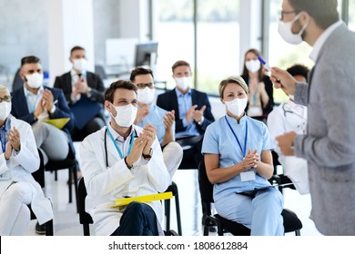 Large group of doctors and business people with protective face masks applauding while attending an educational event at conference hall. Focus is on male doctor. 