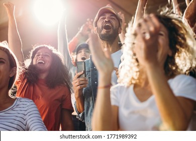 Large Group Of Football Fans In Stadium Yelling And Cheering With Excitement For Their Team. Excited Sports Fans Standing With Their Hands Raised And Celebrating Their Team's Victory.