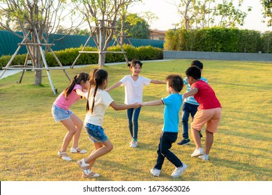 Large group of happy Asian smiling kindergarten kids friends holding hands playing and dancing play roundelay and stand in circle in the park on the green grass on sunny summer day. Multi-ethnic child
