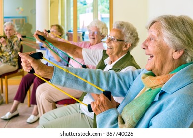 Large group of happy enthusiastic elderly ladies exercising in a gym sitting in chairs doing stretching exercises with rubber bands