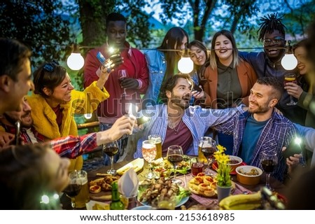 Large group of happy friends singing and having fun during dinner on balcony, mixed age range people celebrating holiday eating together, people hugging each other and using mobile phone lights