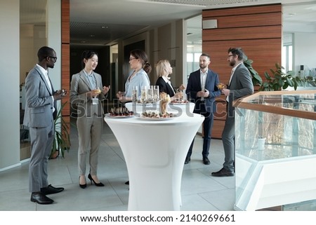 Large group of intercultural people in formalwear communicating at buffet while standing by tables served with snacks and champagne
