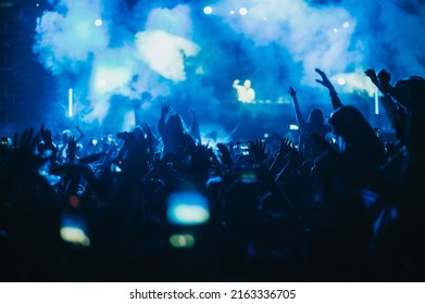 Large Group Of People In A Concert Crowd Enjoying Live Performance On A Music Festival At Night. Audience Raising Hands Up.