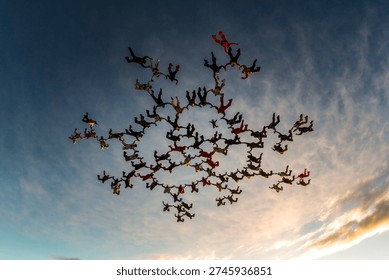 Large group of skydivers creating a complex star formation during freefall at sunset. Aerial teamwork, precision and symmetry captured from below against dramatic sky.