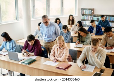 Large Group Of University Students Writing An Exam While Professor Is Supervising Them In The Classroom. 