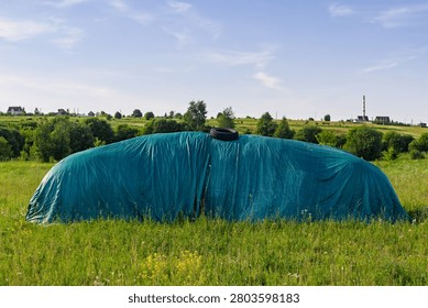 Large haystack covered with a green protective tarp and secured with a tire sits in a lush rural field under a clear blue summer sky, authentic agricultural landscape scenery