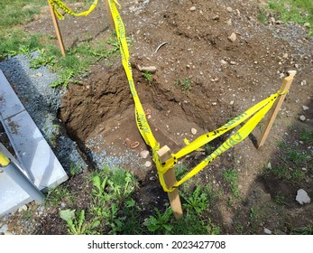 A Large Hole In The Ground With Caution Tape Around It. Two Wooden Posts Are Stuck Into The Muddy Ground To Create A Fence With The Yellow Caution Tape To Warn Pedestrians Of The Potential Fall Hazard