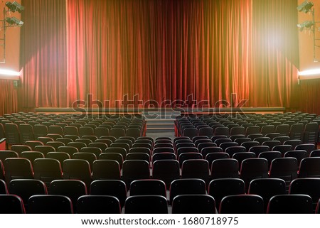 Large illuminated theatre hall with empty seats viewed from the rear looking towards the stage with its closed red curtain in a performing arts concept