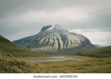 A large, imposing mountain with a snow-capped summit dominates the landscape, surrounded by rolling green hills and a calm body of water.