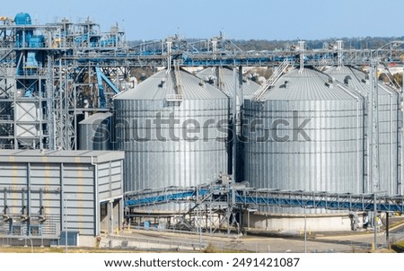 A large industrial facility featuring multiple cylindrical metal silos with conical roofs, surrounded by steel frameworks, conveyor systems, and interconnected pipes. A rectangular building with vertical yellow accents sits in the foreground, while additional industrial structures and a distant landscape of trees and hills extend into the background. The scene provides a detailed view of modern bulk‑storage and processing infrastructure, highlighting the scale and complexity of agricultural and industrial operations.