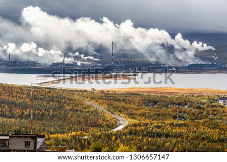 Large industrial metallurgical and chemical plant surrounded by mountains, taiga forest and lakes on a cloudy day. Heavy smoke from smokestacks emit into the air. Monchegorsk, Russia