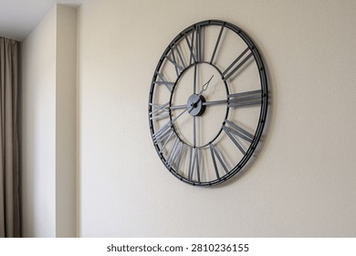large industrial wall clock features a metal frame with oversized 3D Roman numerals and minimalist hands. its dark grey finish contrasts with the pale beige wall