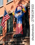A large inflatable Uncle Sam figure stands on a porch in front of a brick building. An American flag waves nearby as the scene captures the excitement of a holiday celebration.