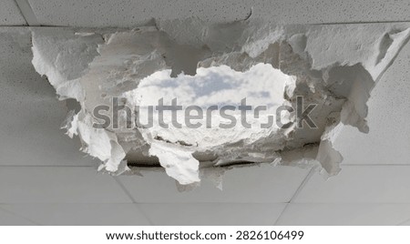 A large, jagged hole in a white ceiling exposes white insulation and a bright sky, indicating structural damage.