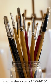 A large jar is filled with paintbrushes inside an artist's studio.