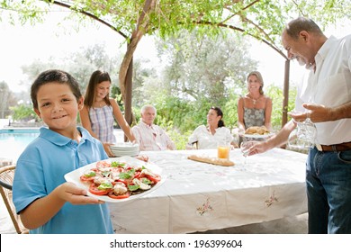 Large Joyful Family Preparing A Table Outdoors Together, Placing Plates, Glasses And Healthy Food During A Sunny Holiday Day In A Vacation Villa Home Garden, Outdoors. Teamwork And Eating Lifestyle.