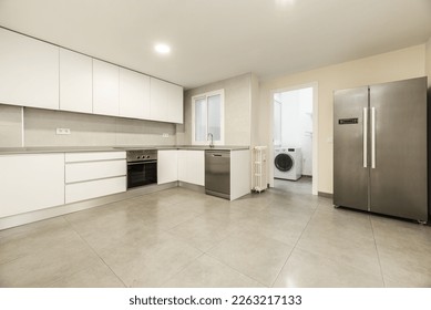 Large kitchen with modern furniture without handles with white doors and drawers, stoneware floors with large gray tiles, appliances and a large stainless steel refrigerator