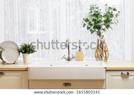 large kitchen sink by the window. a tray with plates, a vase with branches of eucalyptus and cotton, a flower pot with a flower on the countertop.