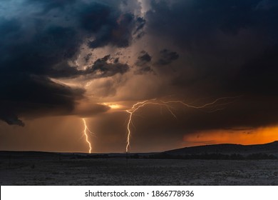 A large lightning strike at dusk in an open plain framed against a deep, dark orange sunset and stormy skies. 