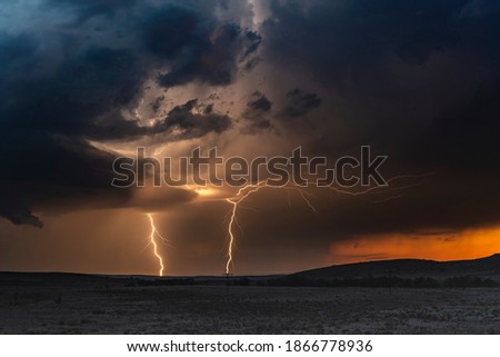 A large lightning strike at dusk in an open plain framed against a deep, dark orange sunset and stormy skies. 
