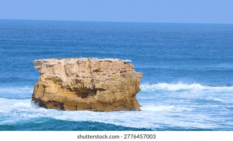 A large limestone sea stack stands amidst blue ocean waves under a clear sky.