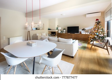 A large living  - dining room with a table, four white eiffel armchairs, a mid century style wooden arm chair, three edison lamps, a three seater sofa and a christmas tree, all on a wooden o