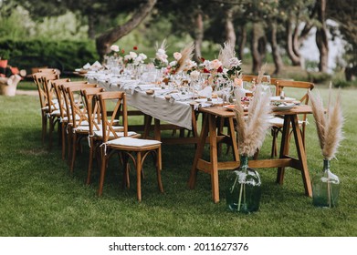 A large, long, decorated, wooden table and chairs, covered with a white tablecloth with dishes, flowers, candles, stands on the green grass in the park, in the forest in nature. Wedding banquet. - Shutterstock ID 2011627376