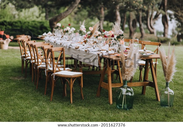A large, long, decorated, wooden table and
chairs, covered with a white tablecloth with dishes, flowers,
candles, stands on the green grass in the park, in the forest in
nature. Wedding banquet.