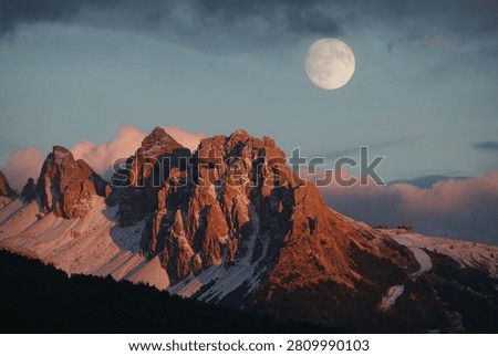 A large, luminous full moon rises above rugged mountain peaks, illuminated dramatically by the golden and warm colors of the sunset or dusk sky.