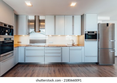 Large Luxury Kitchen Unit Made Of Light Blue Laminate, Glass And Marble. It Is Equipped With Premium Built-in Appliances. The Kitchen Is Lit By Warm Spot LED Lights. The Floor Is Wooden.