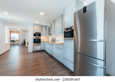 Large Luxury Kitchen Unit Made Of Light Blue Laminate, Glass And Marble. It Is Equipped With Premium Built-in Appliances. The Kitchen Is Lit By Warm Spot LED Lights. The Floor Is Wooden.