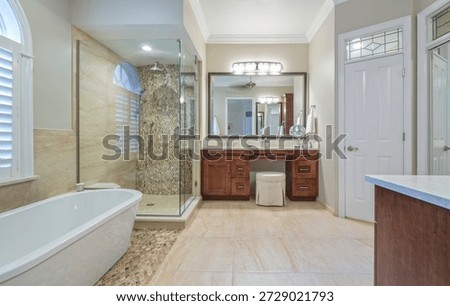 Large master bathroom renovation featuring a white soaking tub and a glass walk in shower with mosaic tile details. The room includes a cherry wood vanity with a large mirror and a white dressing stool. Neutral stone floor tiles and a window with white shutters create a bright airy feel. High end residential interior design with crown molding and modern plumbing fixtures for a luxury home.