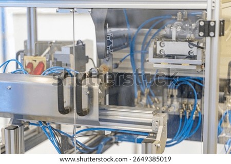 Large metal machine with multiple components, likely precision manufacturing equipment, in an industrial scene Features blue hoses, cables, wiring systems, white connectors, panel with switches, sma