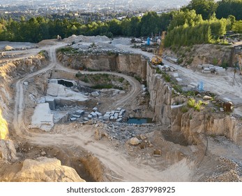 large mining quarry features stepped rock faces and terraced benches, with excavator positioned on upper level. Scattered stone blocks rest among dirt roads, revealing piles and central muddy pool