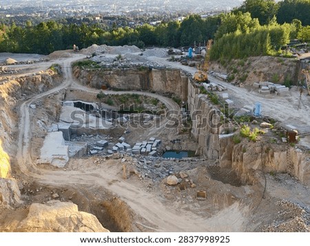large mining quarry features stepped rock faces and terraced benches, with excavator positioned on upper level. Scattered stone blocks rest among dirt roads, revealing piles and central muddy pool