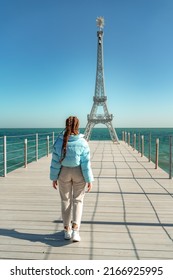 Large model of the Eiffel Tower on the beach. A woman walks along the pier towards the tower, wearing a blue jacket and white jeans.