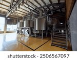 Large, modern brewery with several stainless steel tanks, connected by a network of pipes and valves, with staircase leading to platform above. Concept of beer, brewery, manufacture, quality control