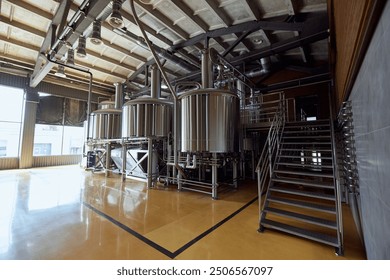 Large, modern brewery with several stainless steel tanks, connected by a network of pipes and valves, with staircase leading to platform above. Concept of beer, brewery, manufacture, quality control - Powered by Shutterstock