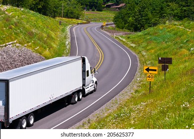 Large Modern Classic White Truck With A Trailer Carrying Commercial Industrial Cargo On The Scenic Highway Winding Among Hills Covered With Trees And Separated The Safety Barrier With Road Signs.