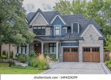 A large modern multi level residential executive home with brown and grey brick stone exterior, shingled roof, beautiful wooden garage doors, interlocked driveway, green gardens and lawn.