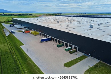 Large modern warehouse with loading docks and parked trucks surrounded by green fields. Industrial logistics hub. Transport infrastructure for distribution, shipping and storage in supply chain system