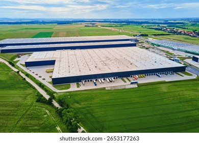 Large modern warehouse with loading docks and parked trucks surrounded by green fields. Industrial logistics hub. Transport infrastructure for distribution, shipping and storage in supply chain system