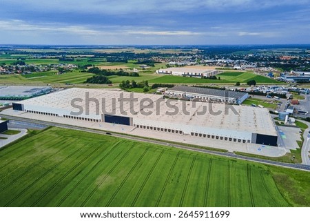 Large modern warehouse with loading docks and parked trucks surrounded by green fields. Industrial logistics hub. Transport infrastructure for distribution, shipping and storage in supply chain system