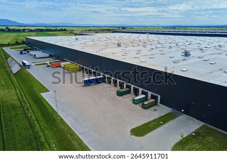 Large modern warehouse with loading docks and parked trucks surrounded by green fields. Industrial logistics hub. Transport infrastructure for distribution, shipping and storage in supply chain system