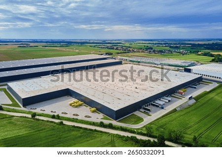 Large modern warehouse with loading docks and parked trucks surrounded by green fields. Industrial logistics hub. Transport infrastructure for distribution, shipping and storage in supply chain system
