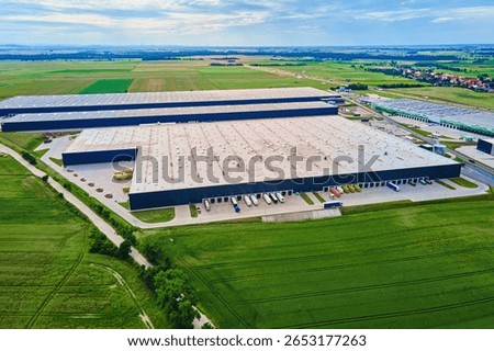 Large modern warehouse with loading docks and parked trucks surrounded by green fields. Industrial logistics hub. Transport infrastructure for distribution, shipping and storage in supply chain system