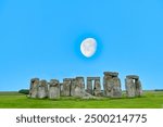 Large moon rising in blue sky above Stonehenge prehistoric structure on Salisbury Plain, Wiltshire, England consisting of outer ring of sarsen standing stones and inside ring of smaller bluestones