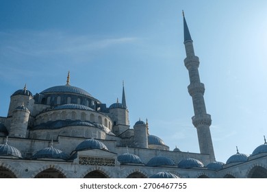 A large mosque featuring multiple domes and a tall minaret under a clear blue sky. The architectural details include rounded structures and a visible tower reaching up. Istanbul, Turkey.