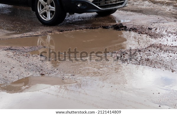 Large muddy puddle on\
broken road on background of front car wheels. Ukraine, Zhytomyr,\
February 9, 2022